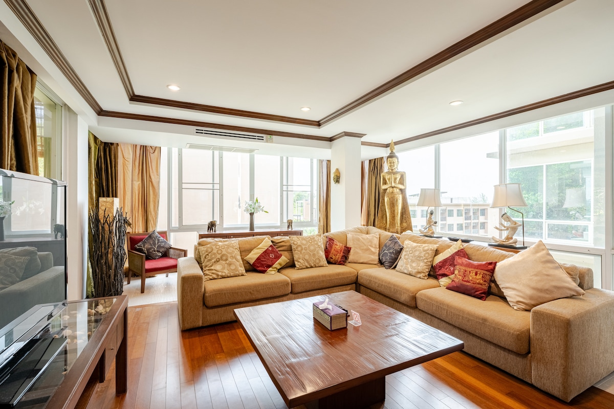 A spacious lounge area features a large, L-shaped sectional sofa adorned with decorative cushions. A wooden coffee table occupies the center, complemented by floor-to-ceiling windows that allow ample natural light. Elegant curtains frame the windows, enhancing the room's ambiance.