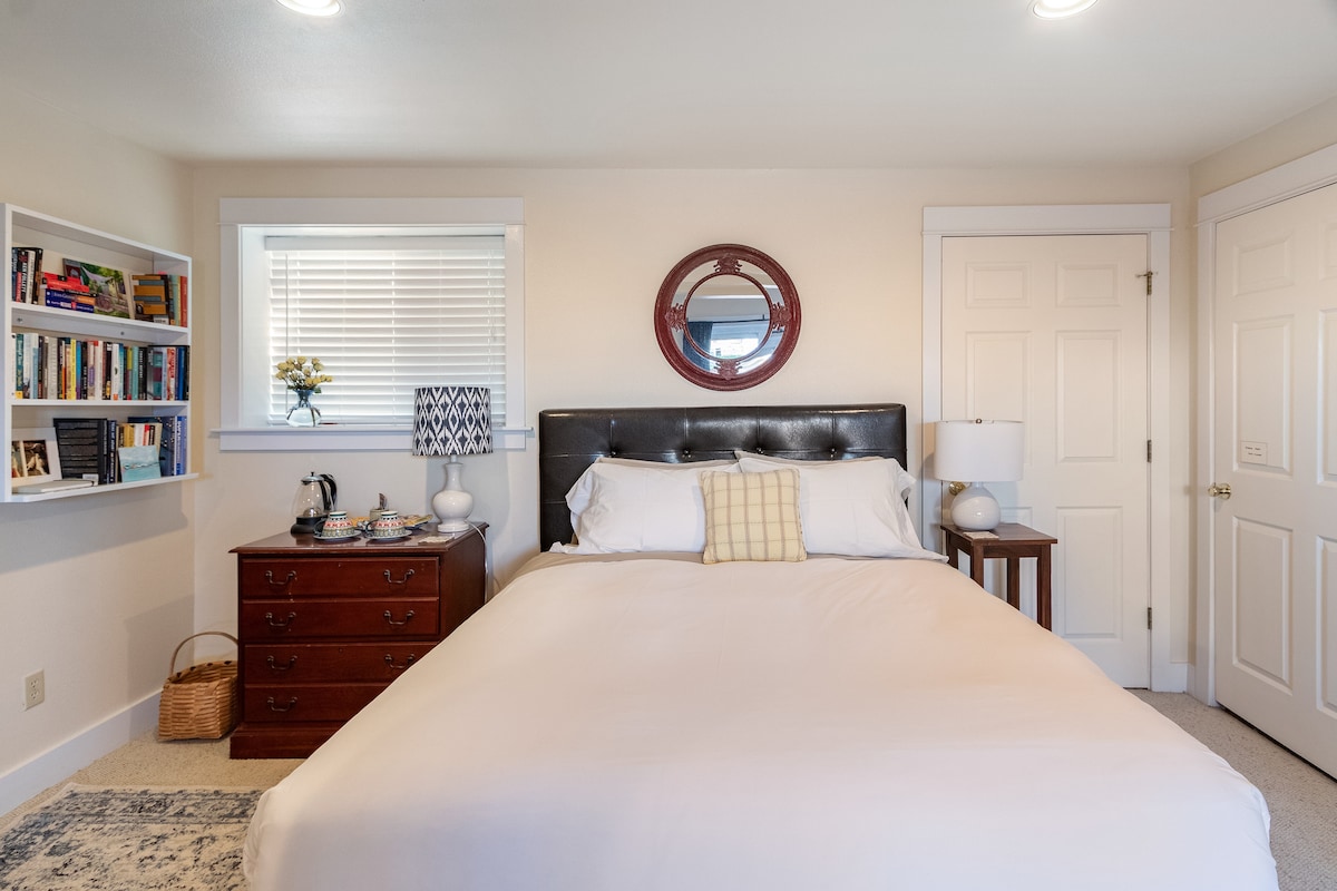 A cozy bedroom features a spacious bed with white bedding and a decorative pillow. A round mirror is mounted above a dark wood dresser, which is accompanied by a bedside lamp and a small basket. Natural light is provided by a window with blinds.