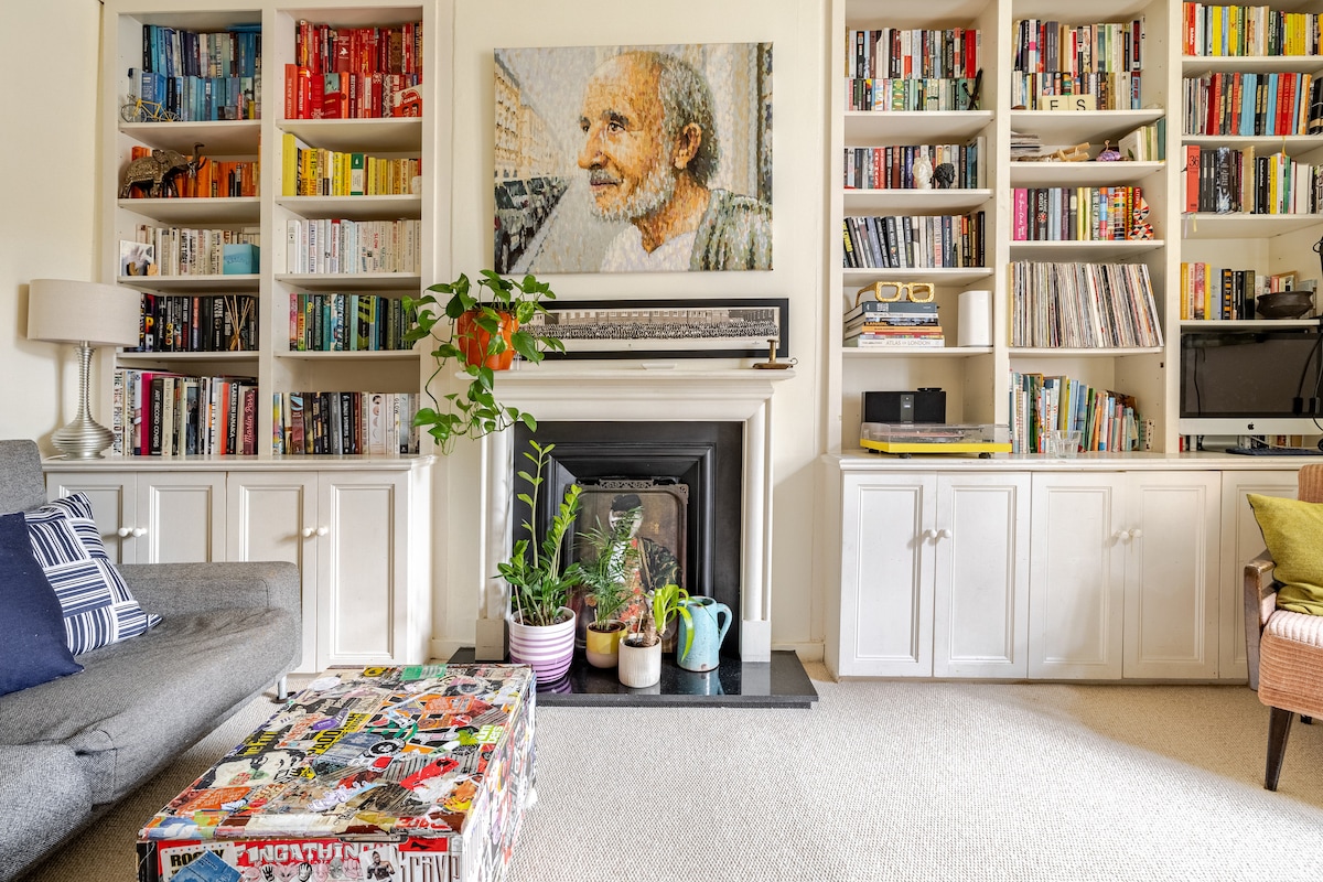 A comfortable living room area is presented, featuring a gray sofa in front of a colorful coffee table adorned with stickers. Bookshelves filled with various titles line the walls, framing a decorative fireplace. A vibrant houseplant adds a touch of greenery to the space.