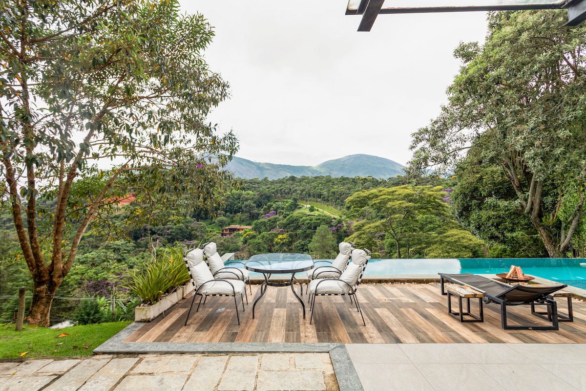 An outdoor terrace showcases a glass table surrounded by six lounge chairs. Lush greenery and mountains form a serene backdrop, while a pool is visible to one side, enhancing the tranquil setting for relaxation.