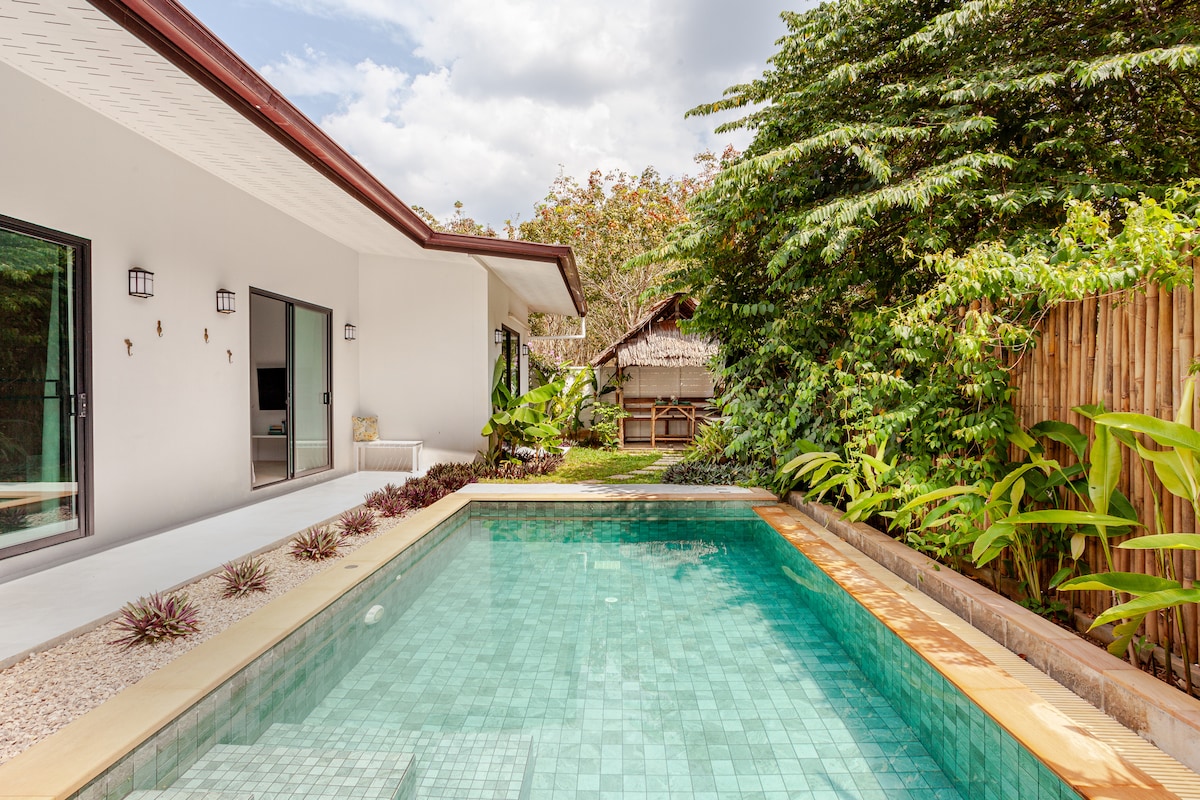 The private plunge pool reflects the surrounding tropical greenery, offering a tranquil space for relaxation. The villa's modern architecture seamlessly integrates with nature, featuring sliding glass doors that provide access to the open-air living area. Lush plants frame the pool area, enhancing the serene environment.