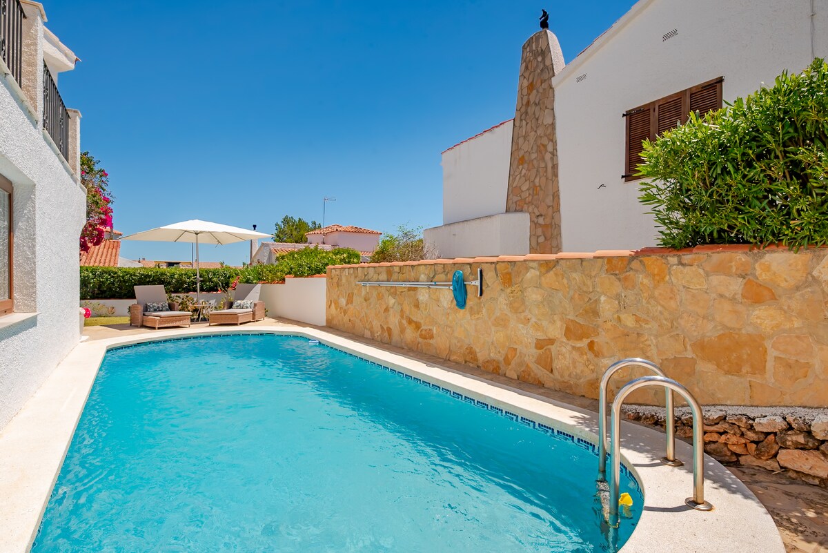 A private pool is surrounded by a stone wall and lush greenery. Two sun loungers and an umbrella are situated next to the pool, creating a relaxing outdoor space. The pool's clear blue water reflects the bright blue sky above.