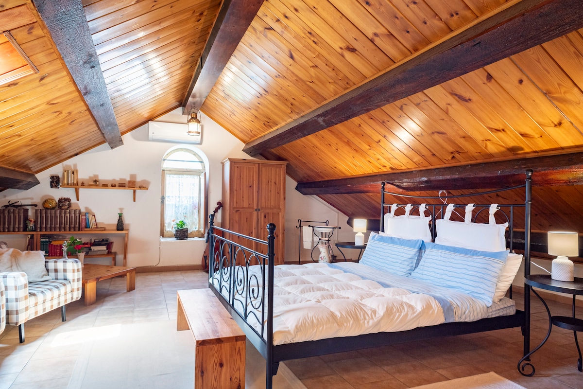 An inviting attic space features a spacious bed with soft white bedding and blue accents. Wooden beams enhance the ceiling's height, while a cozy armchair with a checkered pattern sits nearby. Shelves lined with books and a wooden cabinet provide additional storage.
