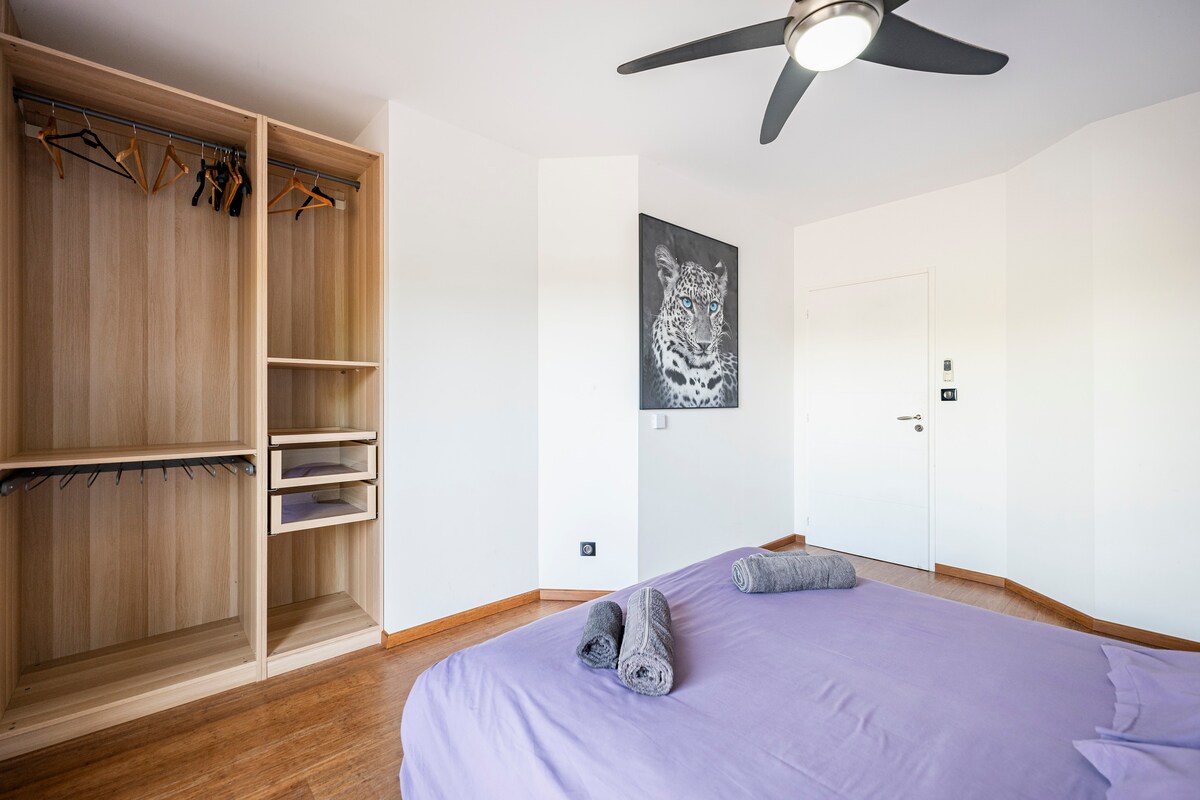 A spacious bedroom is presented featuring a comfortable bed made with purple linens. A modern ceiling fan is installed above, and a wooden wardrobe with hangers is visible. A decorative wall art piece depicting a tiger is showcased, adding a unique touch to the room.