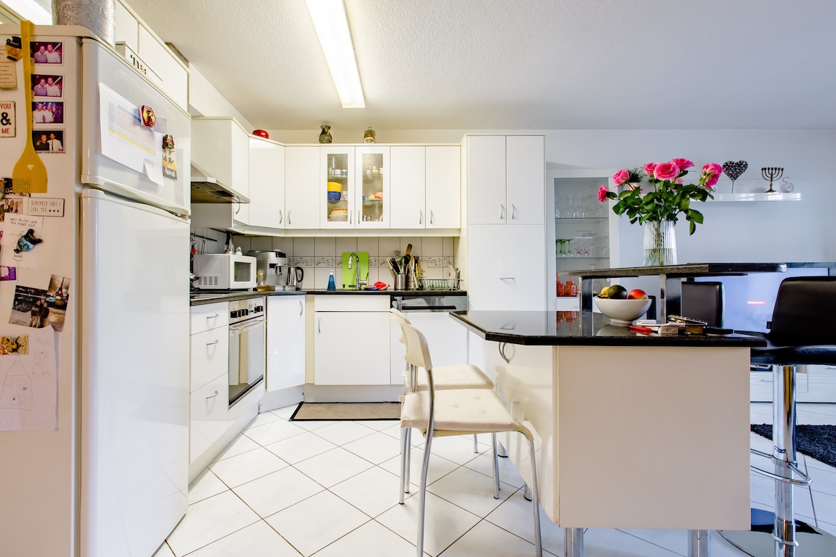 The open kitchen features modern appliances, including a refrigerator and oven, alongside a black countertop. A dining area with a small table and clear chairs is visible. Bright flowers in a vase add a pop of color, while light floods in from windows, illuminating the tiled floor.