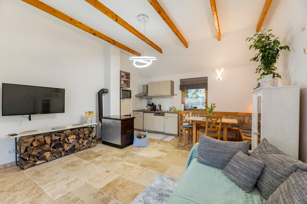 An open living area features a modern kitchen with stylish cabinetry and an inviting dining space. A comfortable seating area includes a light-colored sofa, with a large flat-screen TV mounted on the wall. Natural light highlights the wooden beams overhead.