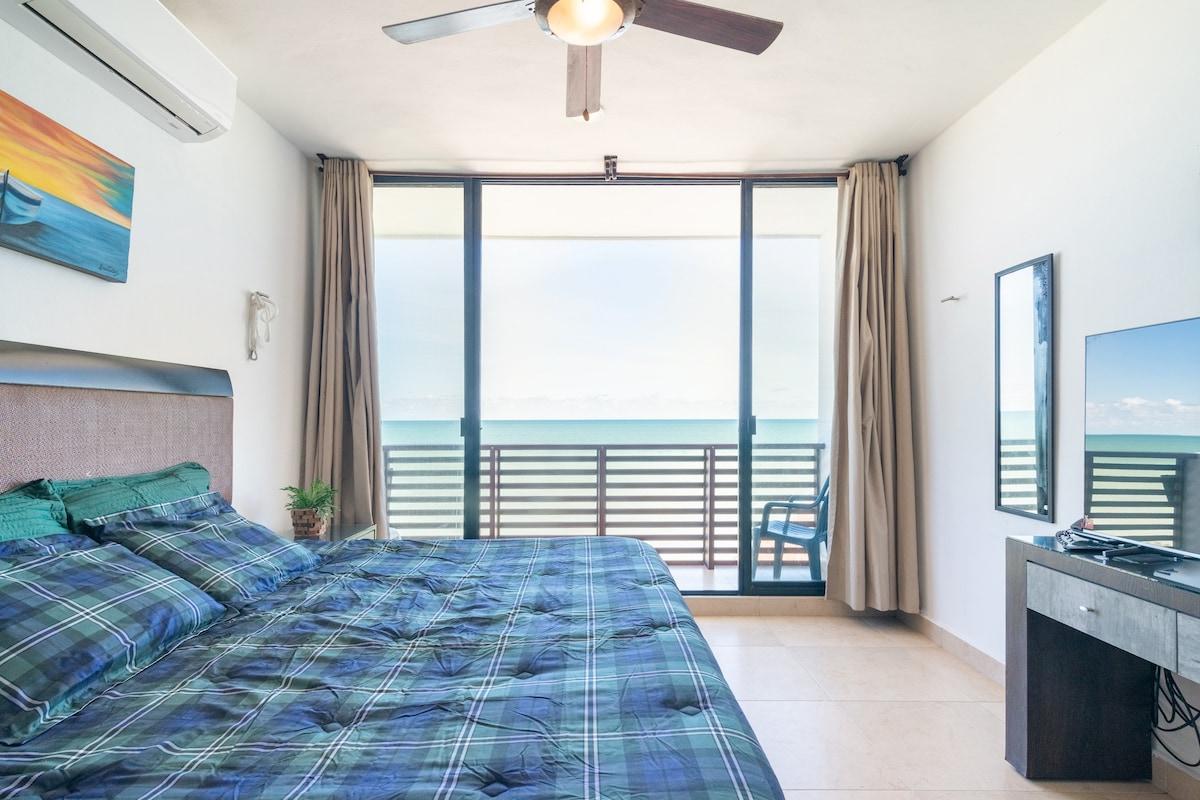 A spacious bedroom features a king-sized bed with a plaid comforter, complemented by blue and green accents. Large glass doors open to a balcony with ocean views. A ceiling fan and air conditioning unit provide comfort, while a flat-screen TV is mounted on the wall.