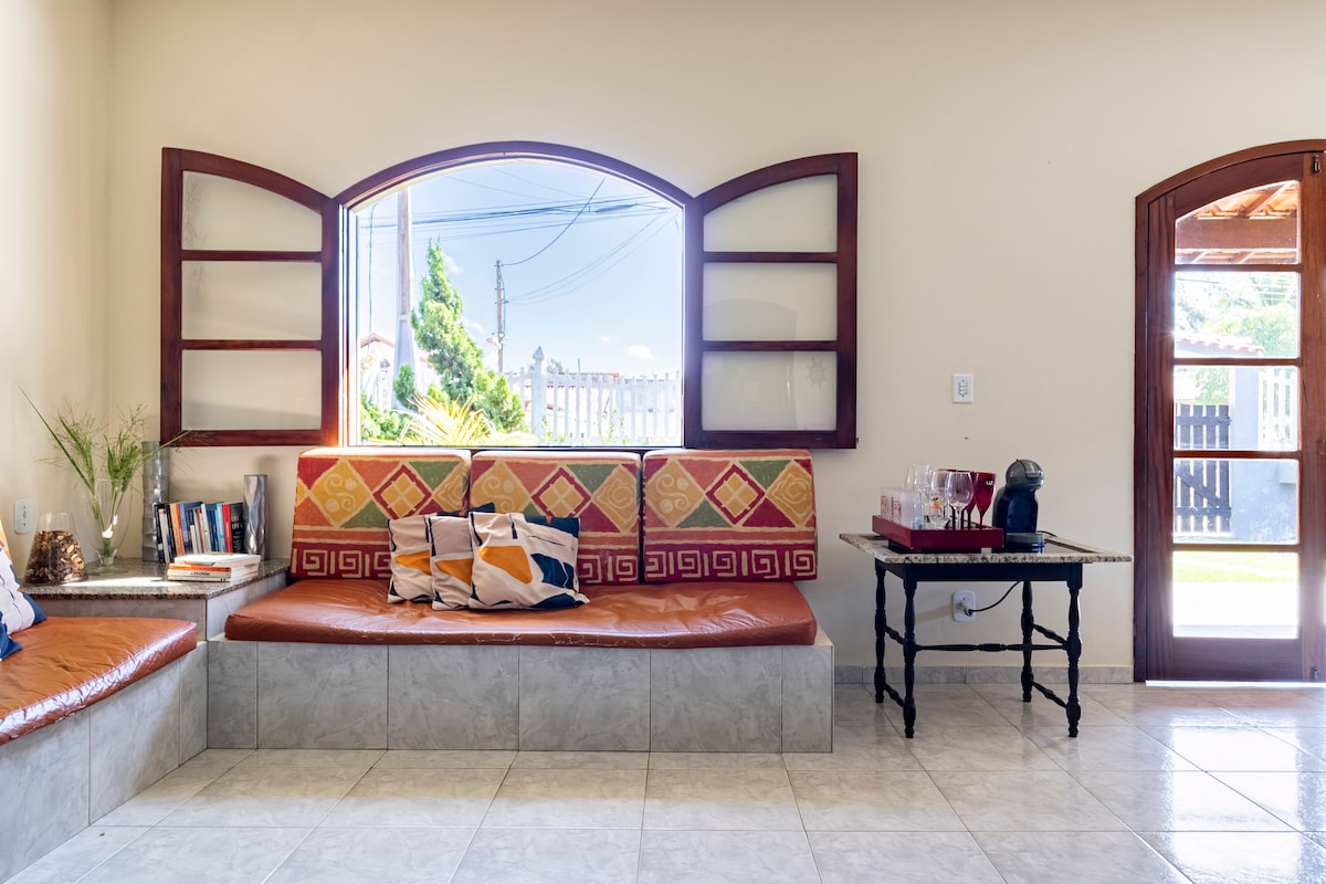 A bright living area features a cozy built-in seating area adorned with colorful cushions. Large windows invite natural light, framing views of the outside greenery. A small table is positioned nearby, enhancing the welcoming ambiance of the space.