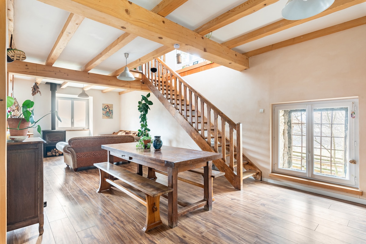 The open living space features a wooden dining table surrounded by ample seating, with large windows providing natural light. A staircase leads to the upper floor, and a cozy seating area is visible, complemented by a wood stove, enhancing the atmosphere of the room.