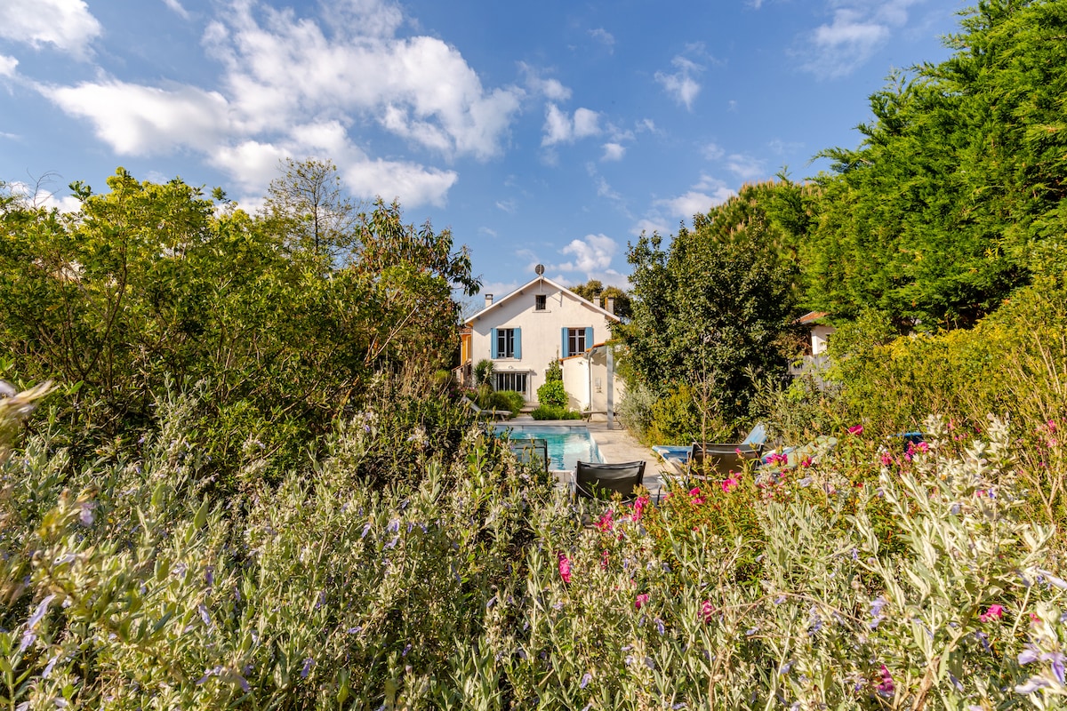 The villa is set within a lush garden filled with blooming flowers and greenery. A spacious pool is visible in the background, with the house prominently featured, showcasing large windows that invite natural light. The serene landscape provides a peaceful ambiance.