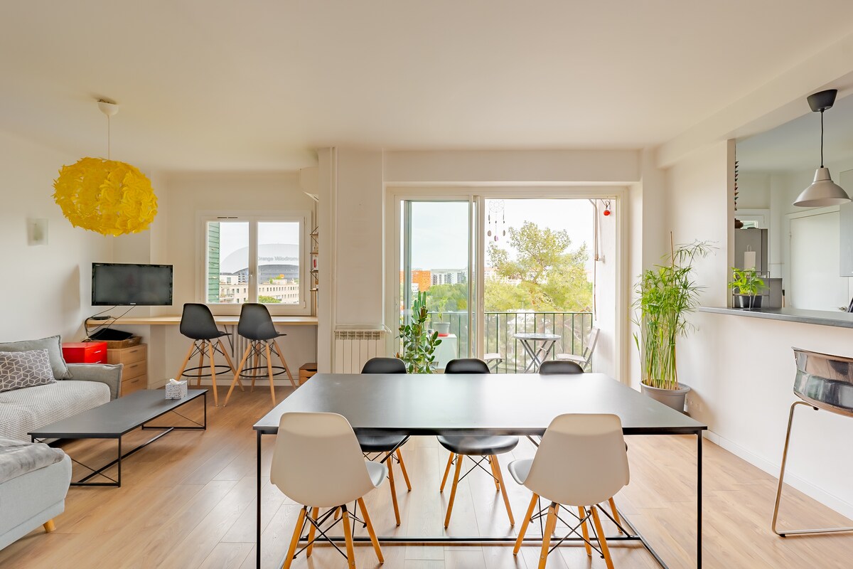 A modern living area is equipped with a dining table surrounded by six chairs, offering a spacious setting. A comfortable sofa is positioned nearby, and large windows provide a view of the outdoor landscape. A decorative light fixture adds a touch of style.