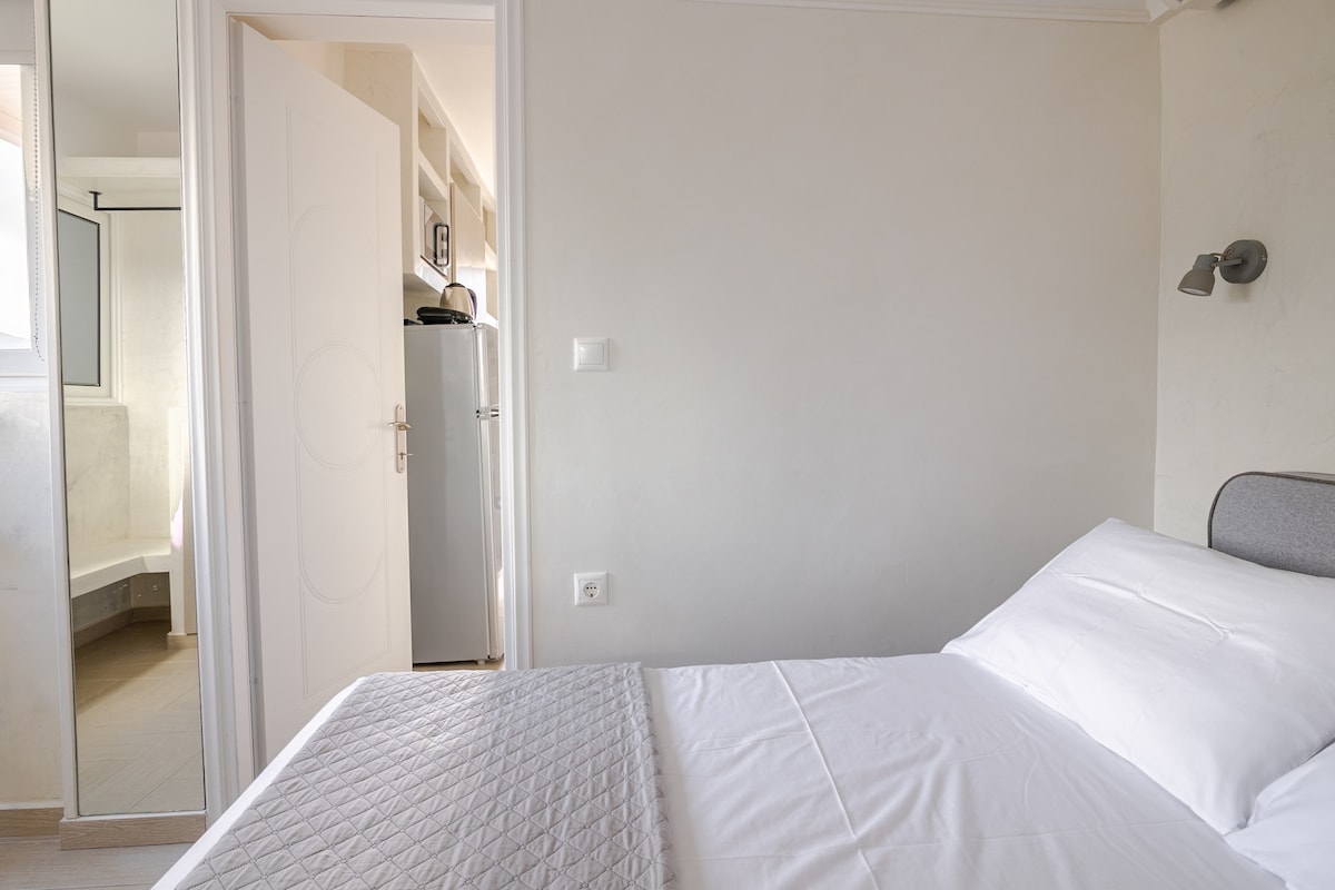 A neatly arranged sleeping area features a spacious bed with crisp white linens. A mirrored wardrobe reflects light, while a door leads towards a modern bathroom. Soft natural light enters through the window, illuminating the neutral-toned walls and contemporary design elements.