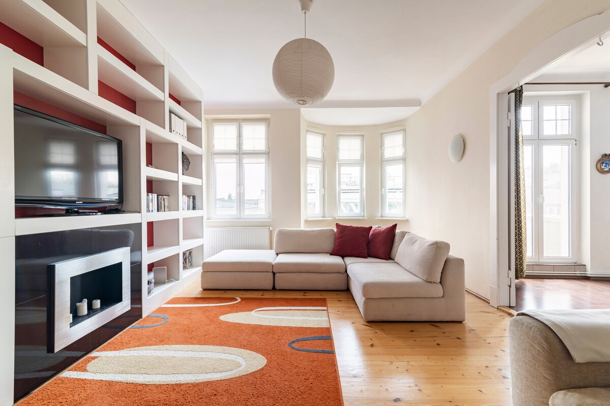 A spacious living area is presented with a light-colored sectional sofa, positioned near a wall-mounted television. Tall windows allow ample natural light to fill the room, highlighting a textured area rug featuring circular patterns. A decorative shelf lines one wall, providing storage and display options.