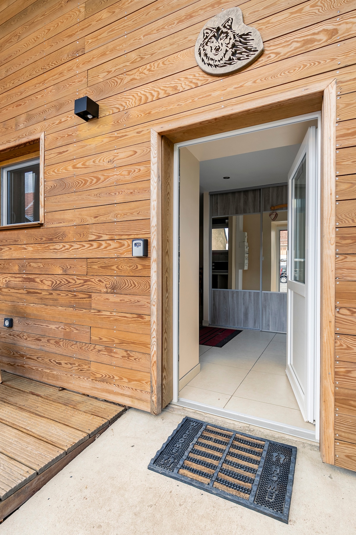 The entrance features a wooden exterior with warm tones, showcasing a decorative wolf sign above the door. A welcome mat is placed at the threshold, leading to a tiled entryway illuminated by natural light.