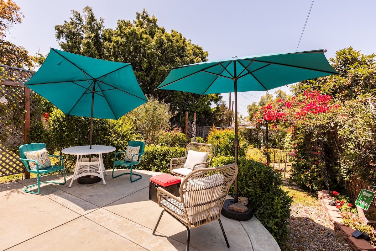 A private garden patio is featured, furnished with a round table and four chairs under large teal umbrellas. Lush greenery surrounds the space, along with vibrant flowering plants, creating an inviting outdoor area for relaxation.