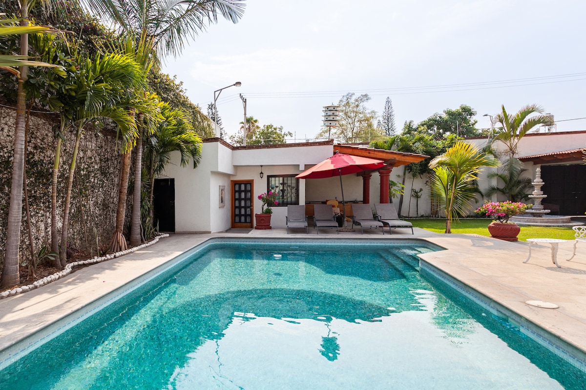 A clear pool reflects the surrounding greenery, with lounge chairs positioned under a large red umbrella. The modern outdoor space features a patio area, complemented by tropical plants and decorative elements, creating a serene environment for relaxation.