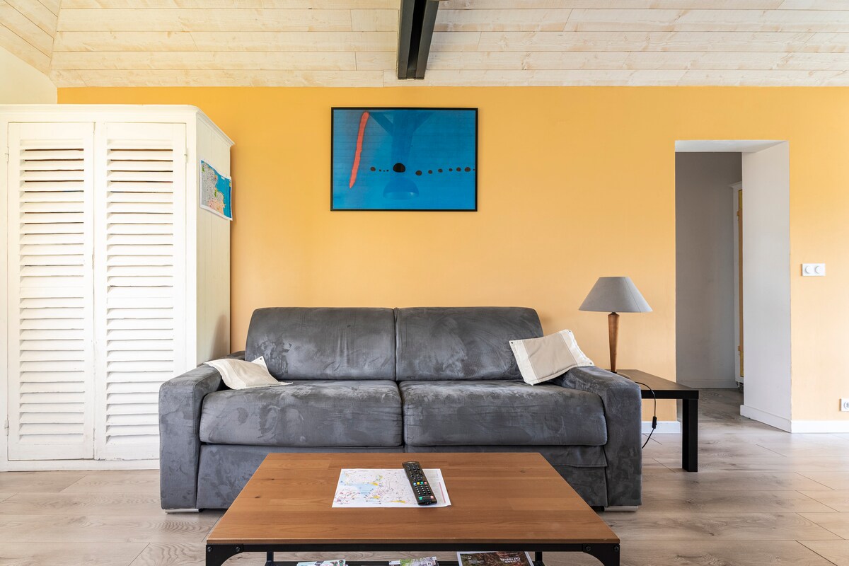 A spacious living area features a gray sofa with decorative pillows, complemented by a wooden coffee table displaying a map. A vibrant artwork hangs above the sofa, while a soft lamp stands nearby. Light-colored walls and wooden accents create a warm ambiance.