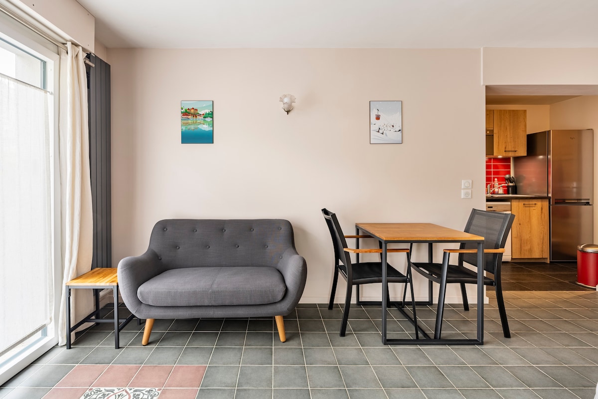 A modern living area features a gray sofa beside a small wooden side table. A dining set comprises a rectangular table and four black chairs. Light-colored walls are adorned with two framed art pieces. Floor-to-ceiling windows allow natural light to fill the space.
