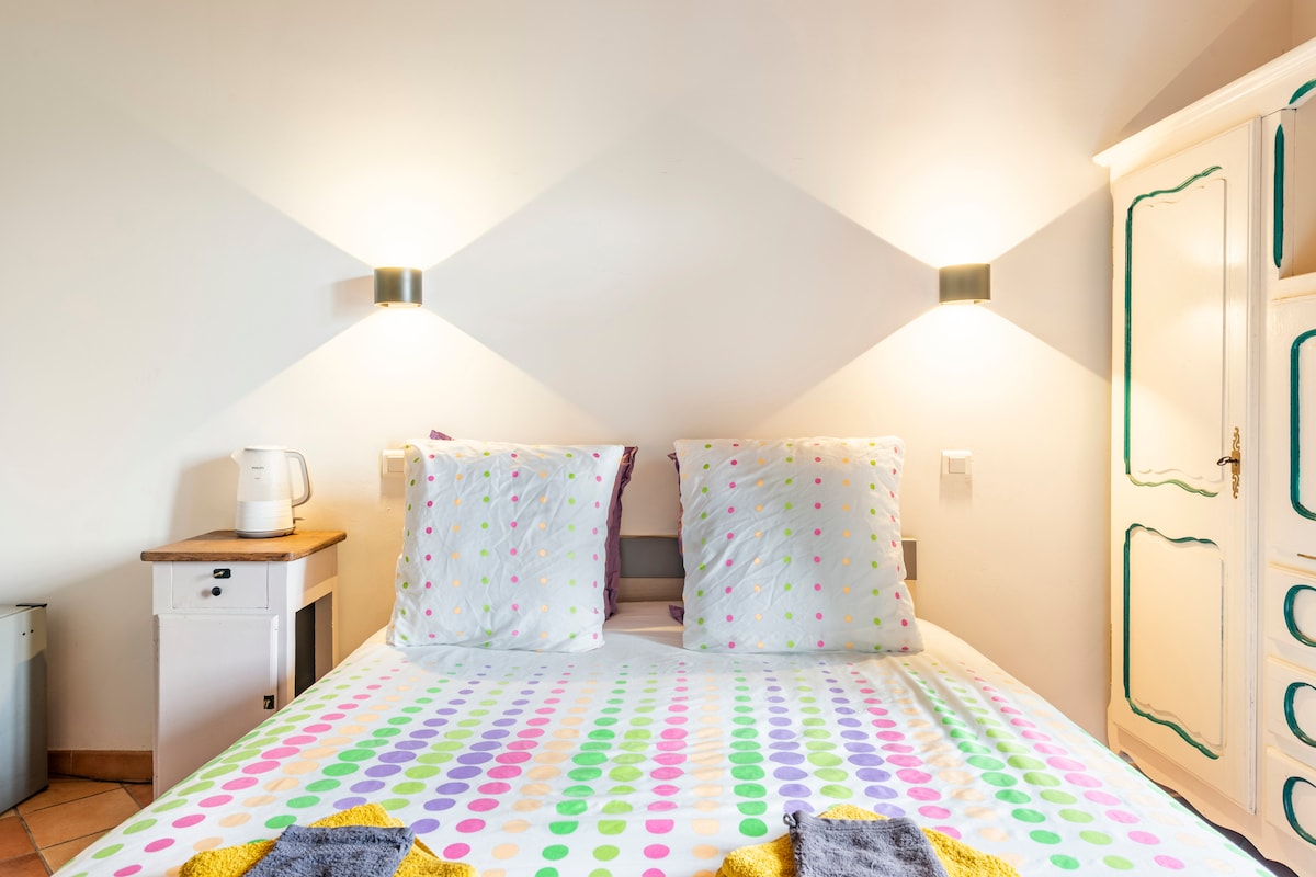 A bright bedroom features a spacious bed with a colorful polka dot duvet cover. Soft lighting is provided by wall sconces on either side of the bed. A small bedside table holds a kettle, while an ornate cabinet adds character to the room.