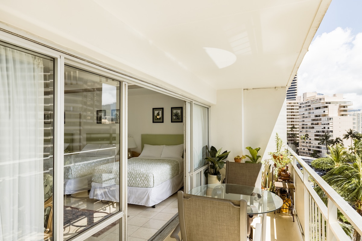 A bright and airy balcony area offers a glass table alongside two chairs, surrounded by tropical plants. The interior features a spacious bed with soft bedding and framed artwork. Large sliding glass doors provide an open view of the outside, enhancing the light-filled ambiance.