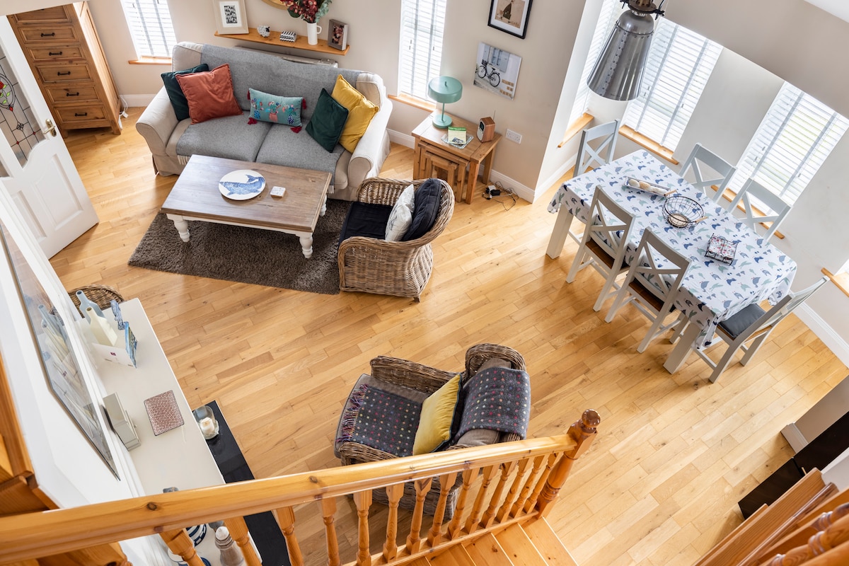 The open-plan living area features a comfortable gray sofa adorned with colorful cushions, accompanied by a stylish coffee table on a soft area rug. A dining table with light-colored chairs sits nearby, while natural light fills the room through white shutters.