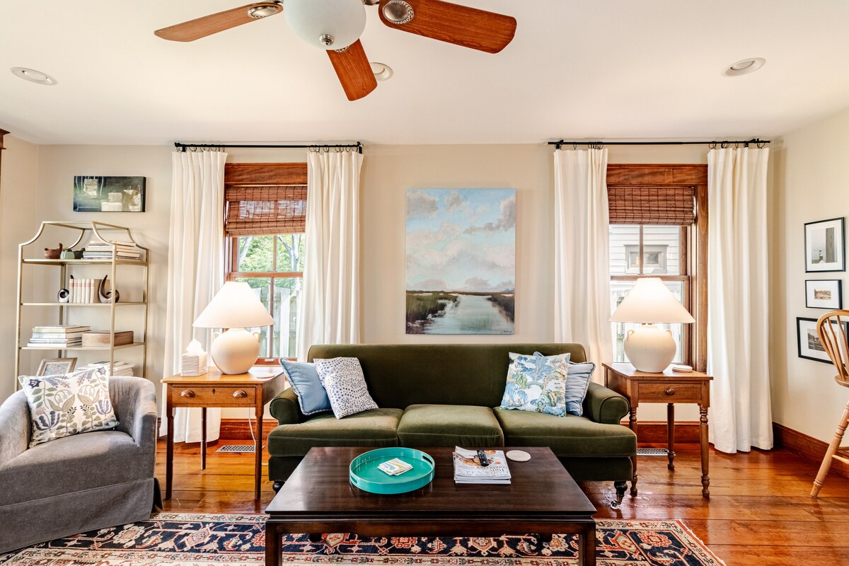 A living room is showcased with a green sofa adorned with throw pillows. A wooden coffee table sits in front, complemented by two side tables with lamps. Windows draped with light curtains allow natural light to brighten the space, enhanced by a ceiling fan.