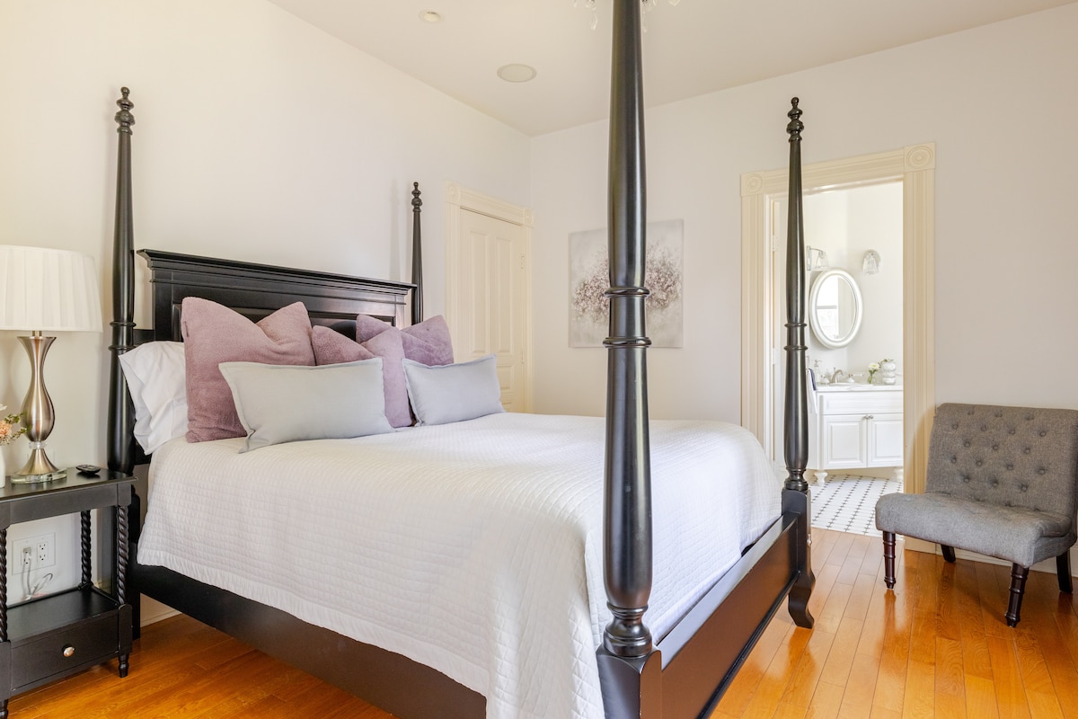 A well-appointed bedroom features a four-poster bed with a light-colored quilt, accentuated by plush lavender pillows. A comfortable armchair sits beside the bed, while a doorway provides a view into an en-suite bathroom adorned with white fixtures.