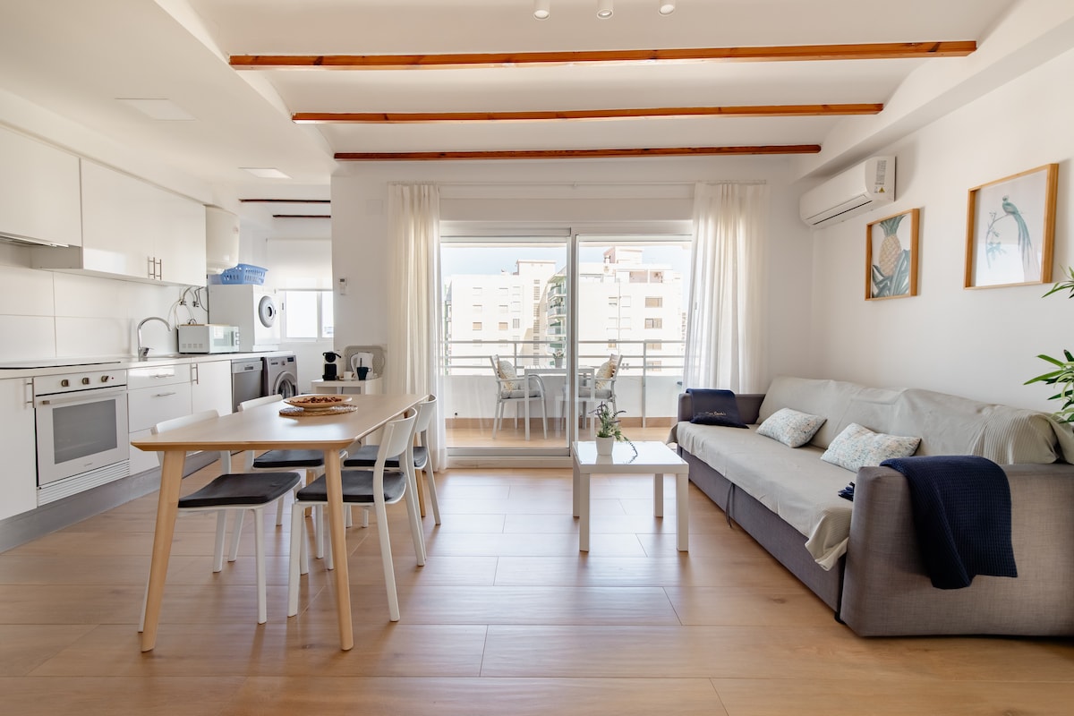 A bright living space is observed, featuring a comfortable sofa, a dining table with chairs, and large sliding glass doors that lead to a balcony. The kitchen is visible in the background, equipped with modern appliances, and decorative wall art adds a touch of warmth.
