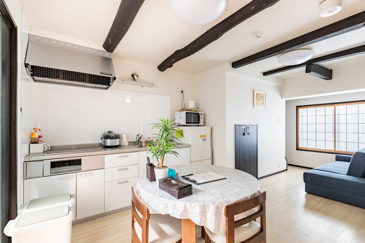 A bright kitchen and dining area features natural light from a large window. A round table with wooden chairs is positioned adjacent to modern kitchen appliances, including a stove and microwave. A comfortable seating area with a blue sofa is visible alongside decorative plants.