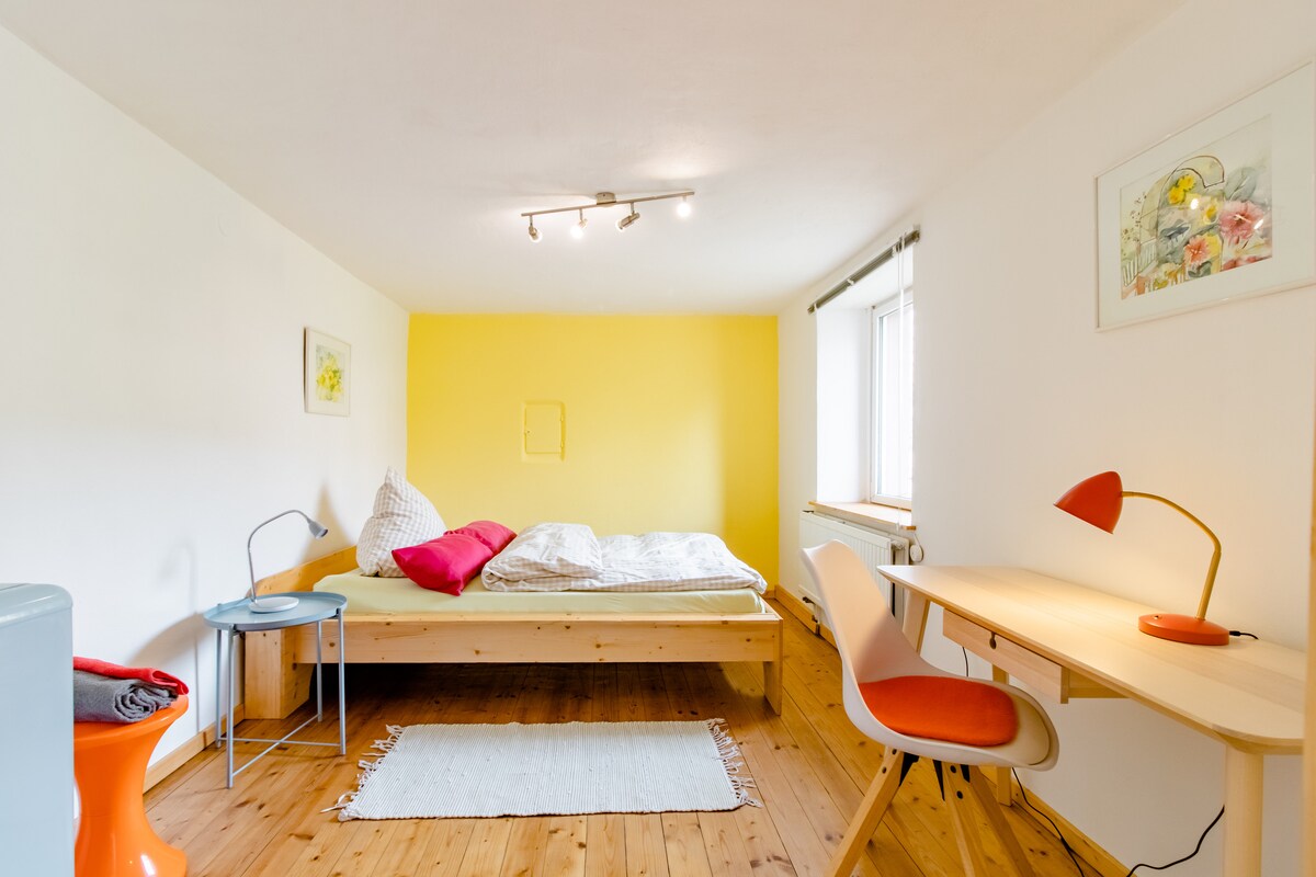 A cozy room features a wooden bed with soft bedding, complemented by a bright yellow accent wall. A small desk with a lamp and an ergonomic chair provide a workspace. Natural light enters through a window, illuminating the space. Pine floorboards add a warm touch.