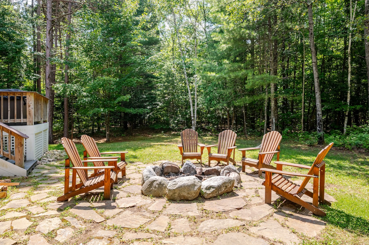An outdoor fire pit area is surrounded by a circle of five wooden Adirondack chairs, set on a stone patio. The backdrop features lush greenery from the surrounding trees, creating a serene and inviting space for gatherings.