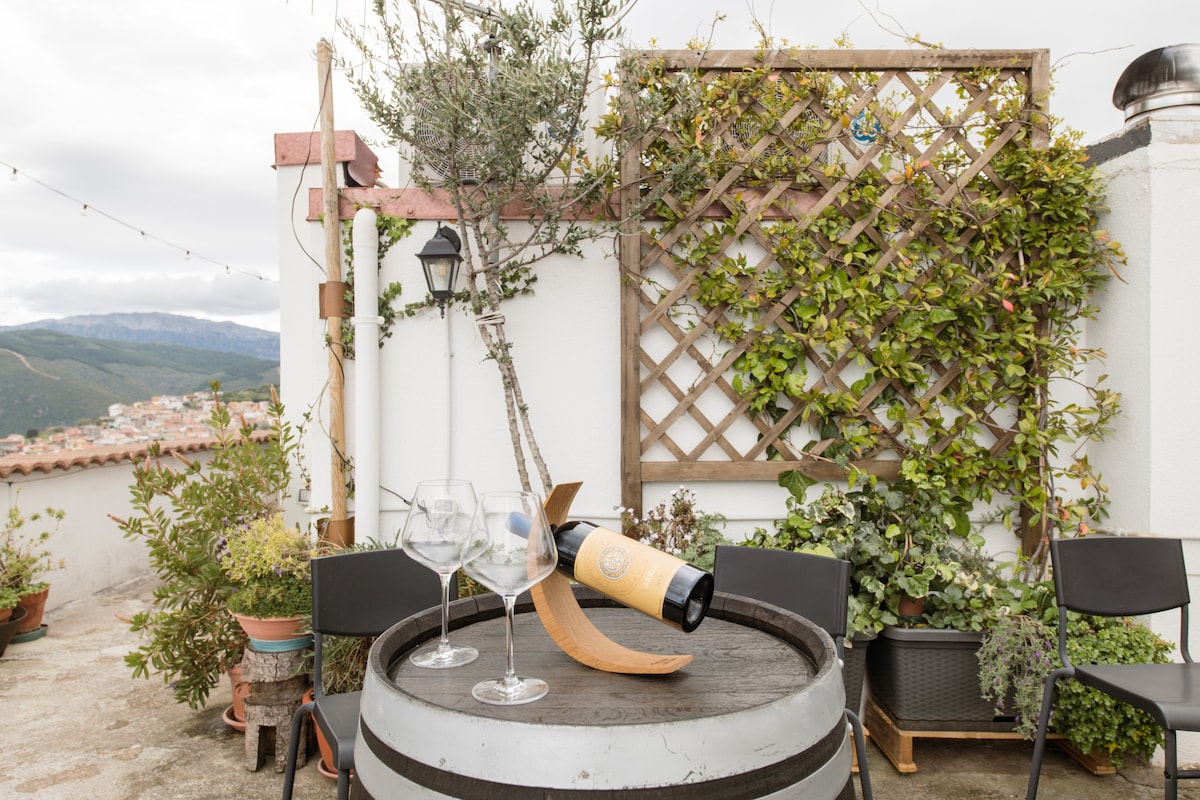 A charming outdoor area is depicted, featuring a wooden wine barrel table supporting a wine bottle and two glasses. Lush greenery surrounds the space, with climbing plants on a trellis in the background, creating a serene environment.