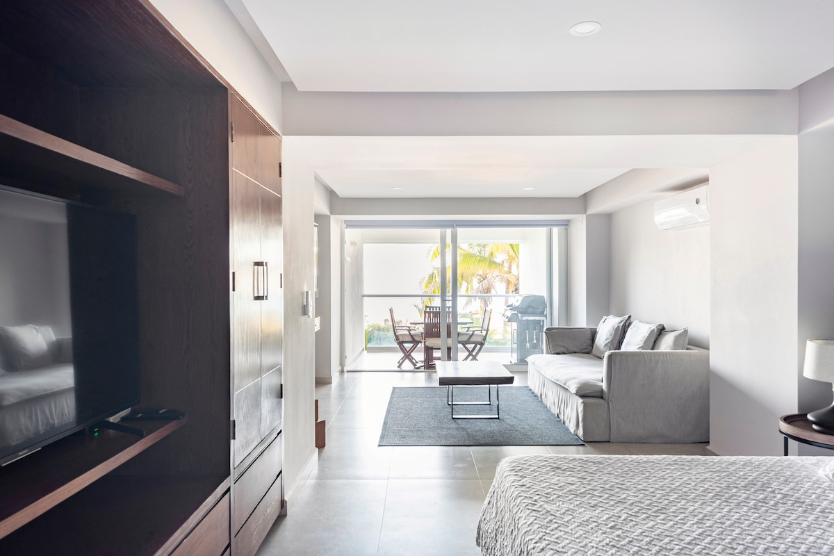 A modern studio layout is presented, featuring a spacious bed and a comfortable seating area with a gray sofa. A glass sliding door offers natural light and views of the outdoor area. A small dining table is visible, enhancing the functional space.