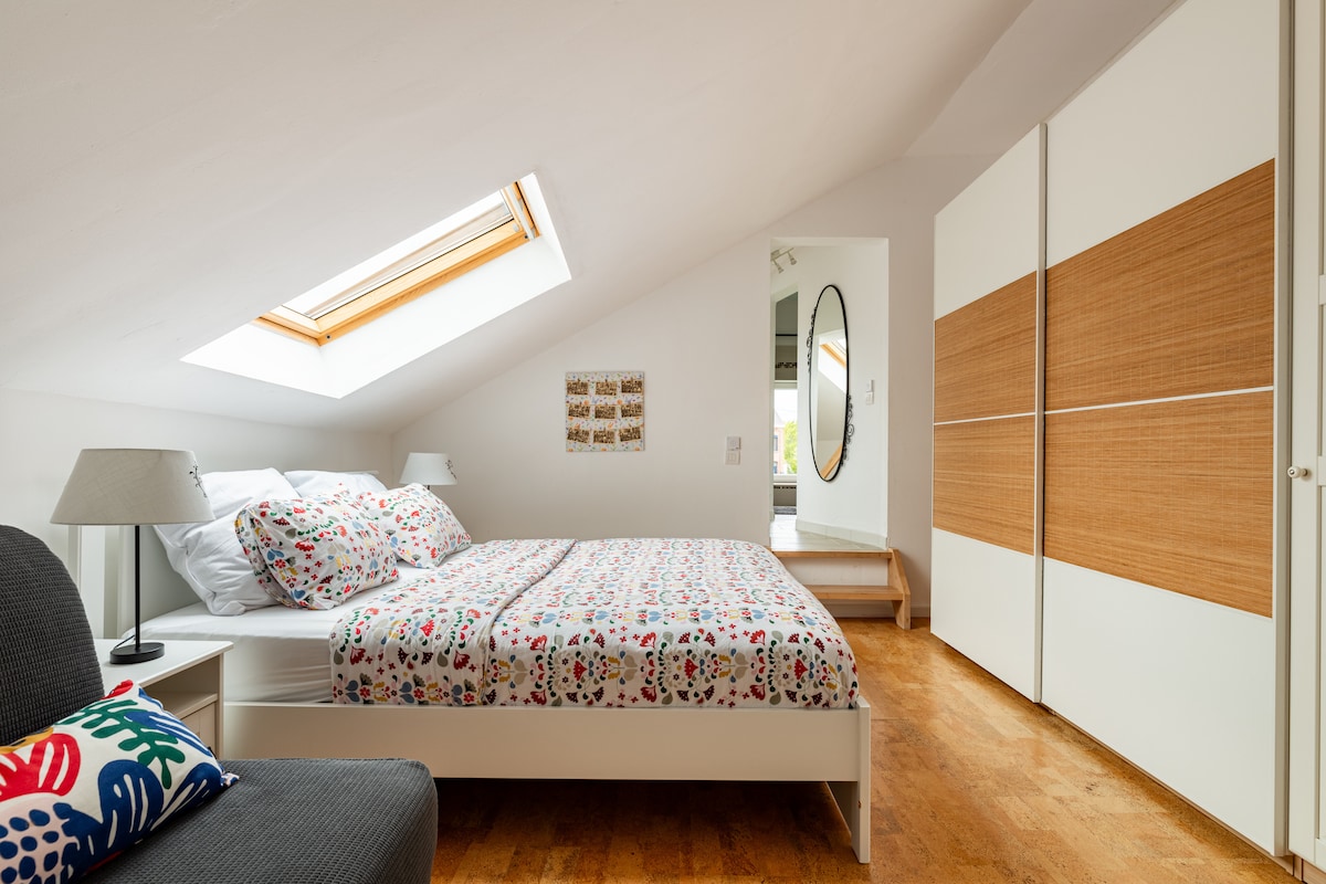 A bright sleeping area is illuminated by natural light streaming through a skylight. A spacious bed with a colorful patterned duvet sits against a white wall. A mirror and wooden storage unit are visible, adding to the room's modern decor.
