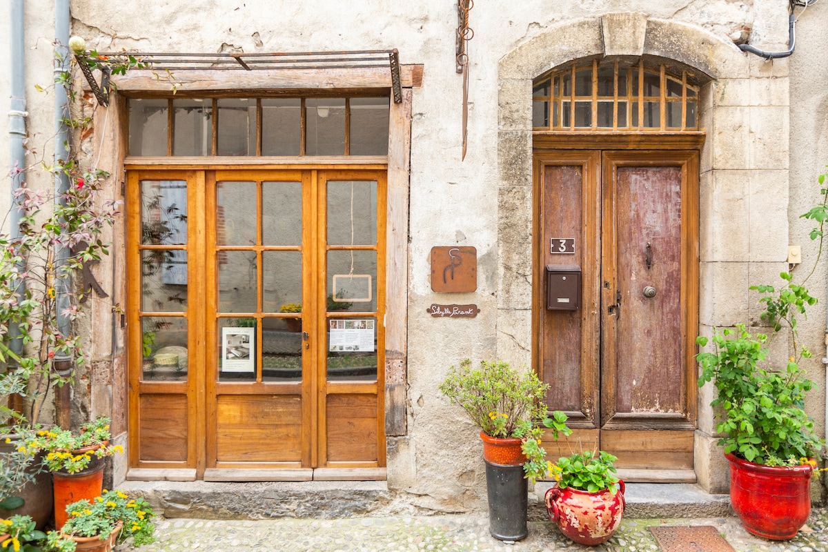 The entrance showcases two wooden doors, one with large windows, adorned with greenery in terracotta pots. A sign hangs on the wall, and decorative elements enhance the charm of the rustic facade, inviting guests into the vibrant atmosphere of the area.