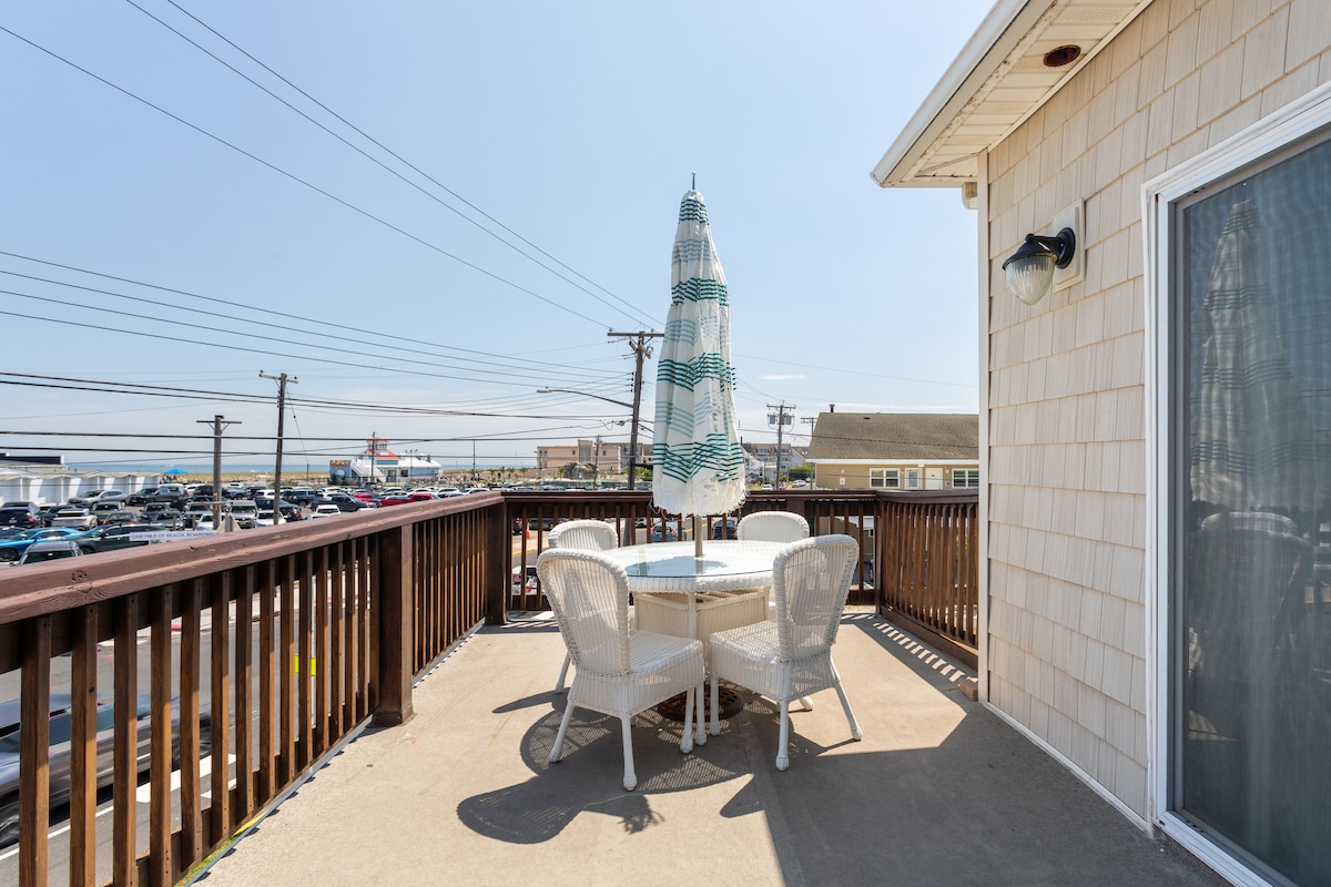 A private deck features a dining table surrounded by four chairs, shaded by a large umbrella. The open sky and distant view of the beach can be seen beyond the railing, creating a relaxed outdoor setting for dining or gathering.
