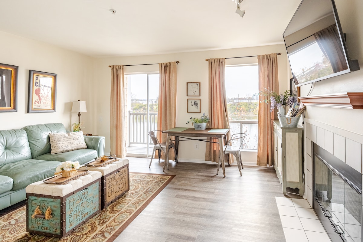A living area features a light green couch with decorative pillows and two rustic coffee tables. A dining area with a small table is positioned by large windows that allow natural light to filter in. Soft curtains frame the windows, and a fireplace is present for added comfort.