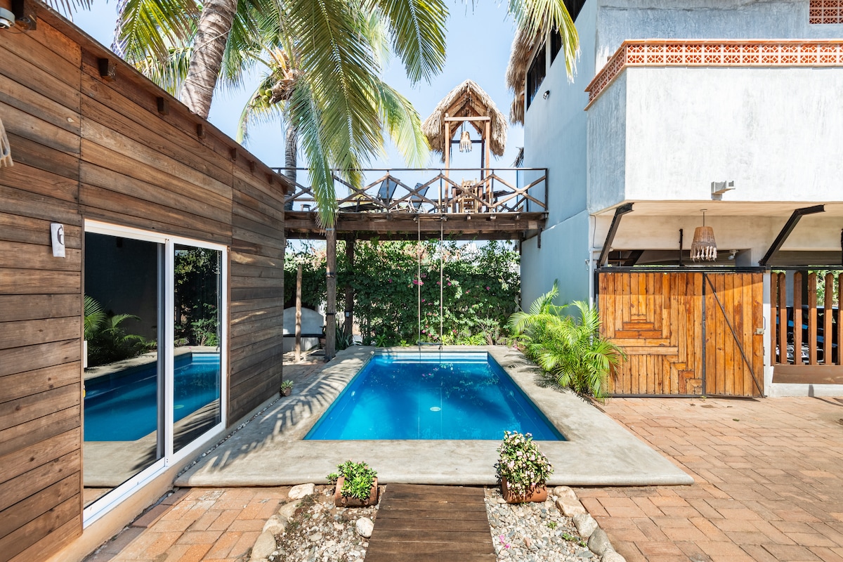A serene swimming pool is surrounded by tropical greenery, with a wooden deck and a shaded pergola above. The exterior walls are a combination of natural wood and stucco. A pathway made of stone leads toward the inviting pool area.