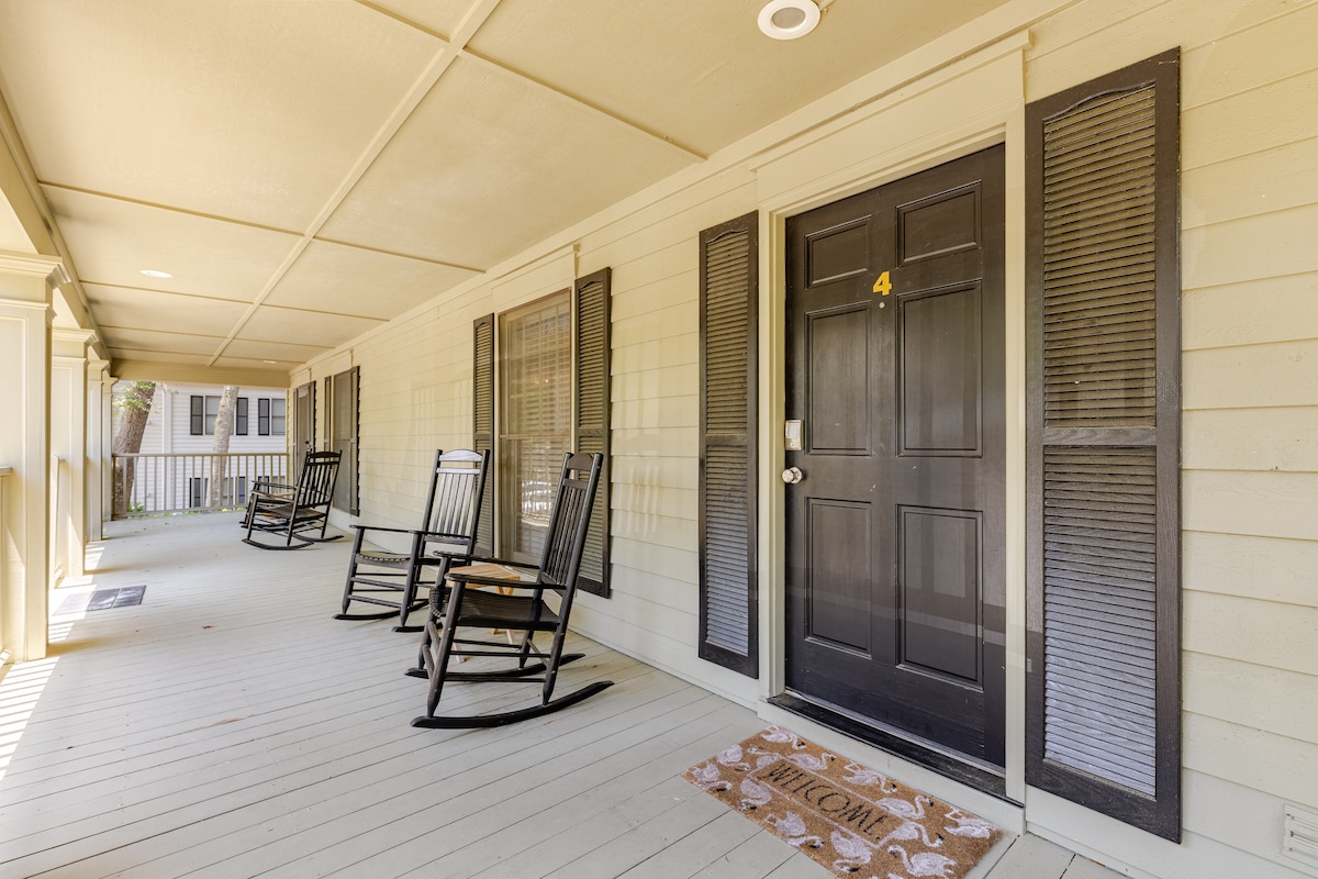 A spacious front porch is furnished with two rocking chairs positioned along the wall. The entrance door is framed by tall windows, allowing natural light to filter in. A welcome mat lies at the foot of the door, creating a friendly first impression.