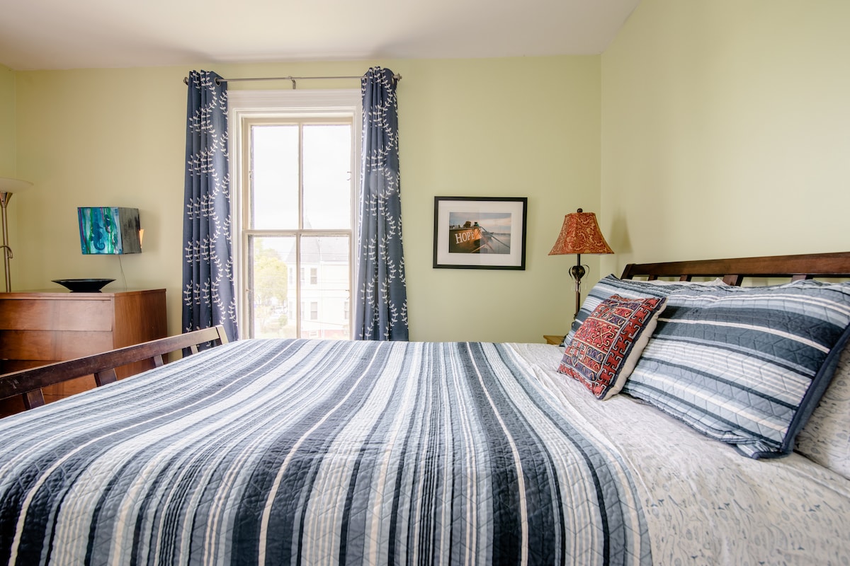 A private bedroom features a spacious bed adorned with striped bedding and decorative pillows. A bedside lamp and framed wall art add character, while large windows are dressed with patterned curtains, allowing natural light to fill the room. A chest of drawers complements the warm decor.