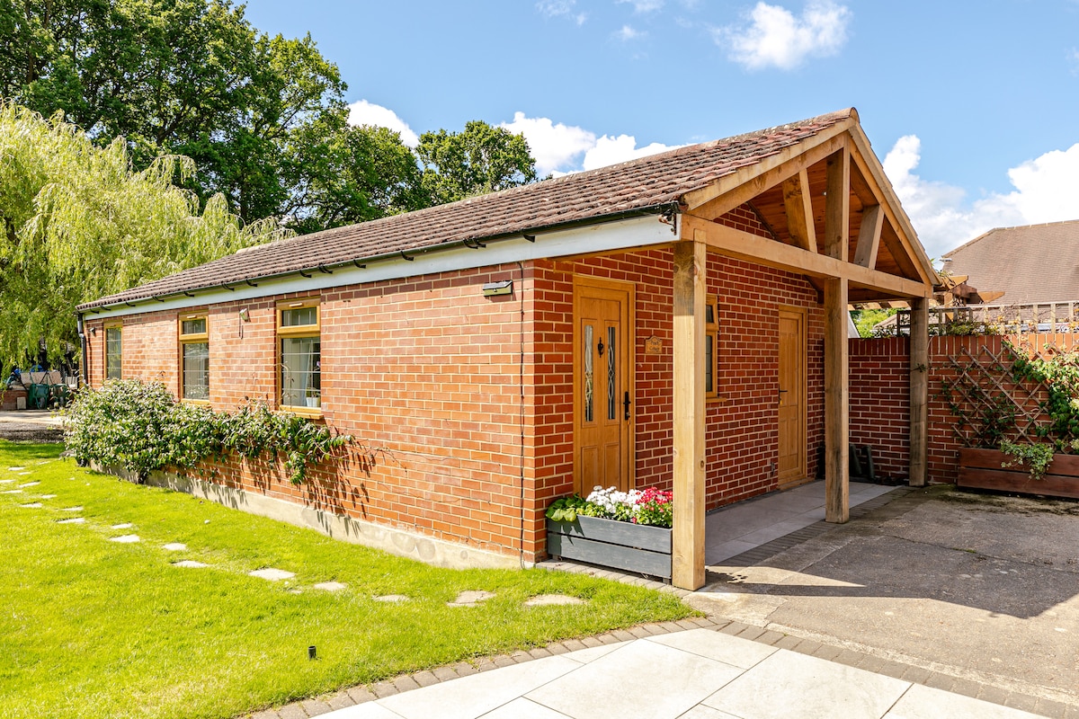 The cottage exterior is characterized by a combination of red brick walls and a wooden overhang. Lush green grass surrounds the entrance, framed by flowering planters. A pathway of stepping stones leads to the covered entrance, providing a welcoming access point.