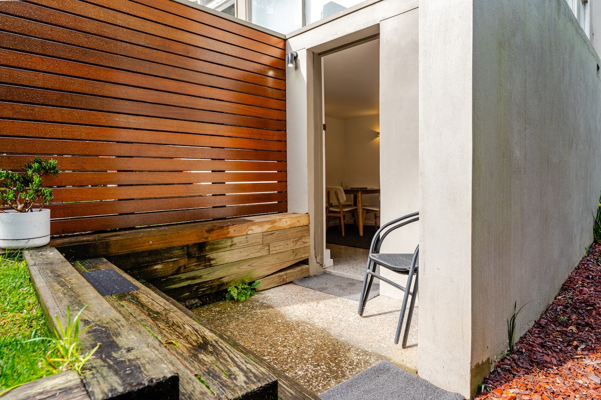 An entrance to a contemporary unit is presented, featuring a clean pathway leading to an open door. Wooden screens provide privacy, while a chair and rustic wooden seating area are positioned to enhance outdoor relaxation.
