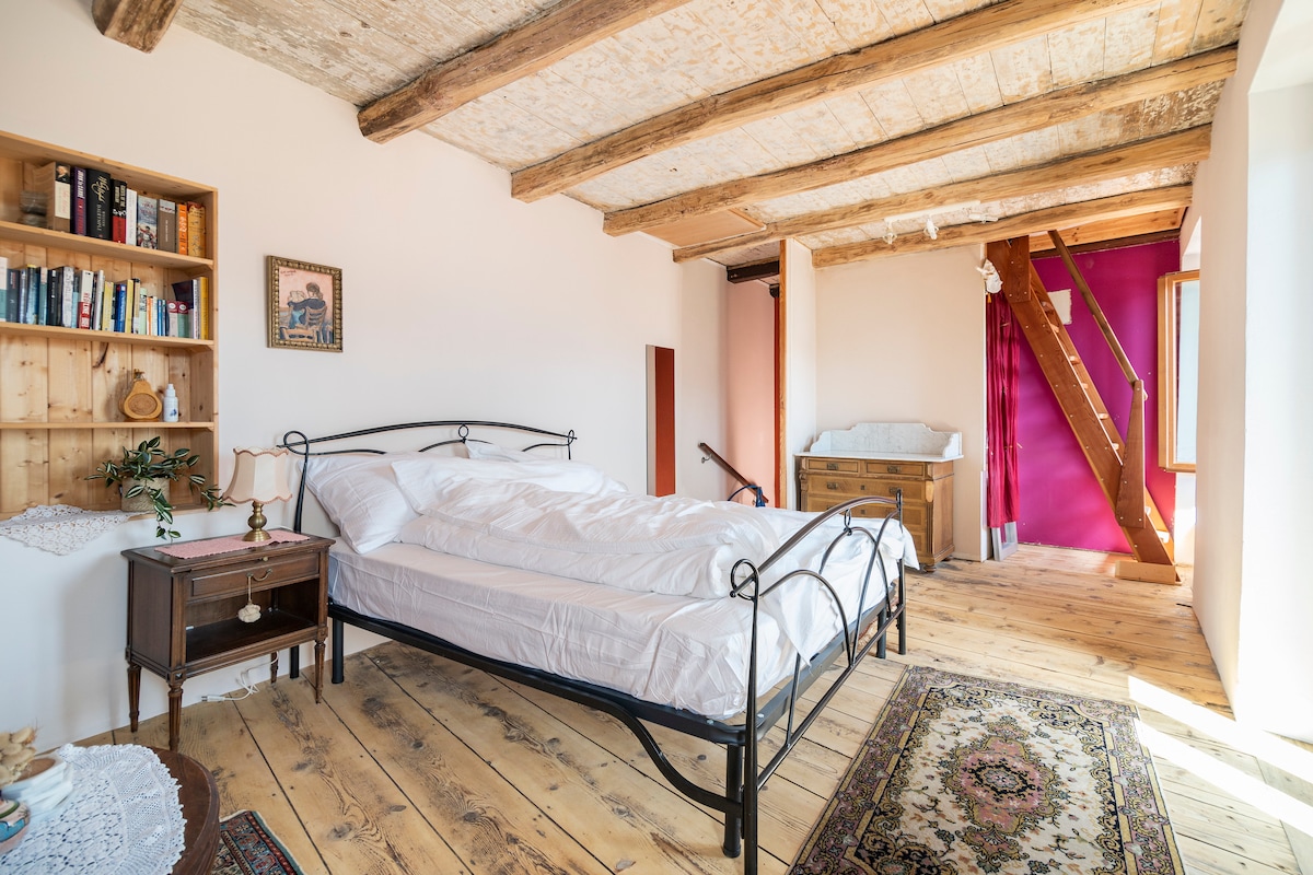 A spacious bedroom features a black metal bed with white bedding, complemented by wooden flooring and exposed wooden beams. A small bookshelf is positioned against the wall, and a dresser is visible in the background. Natural light fills the room through a large window.
