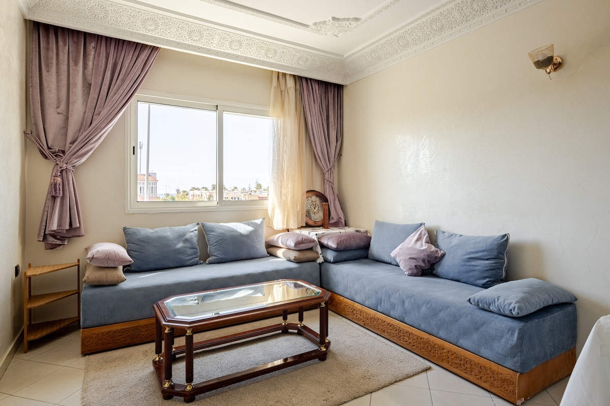 A spacious living area is furnished with a large, L-shaped couch, adorned with soft cushions. A glass-topped coffee table sits in the center, reflecting natural light from a window draped with soft curtains, while decorative elements enhance the room's comfort.