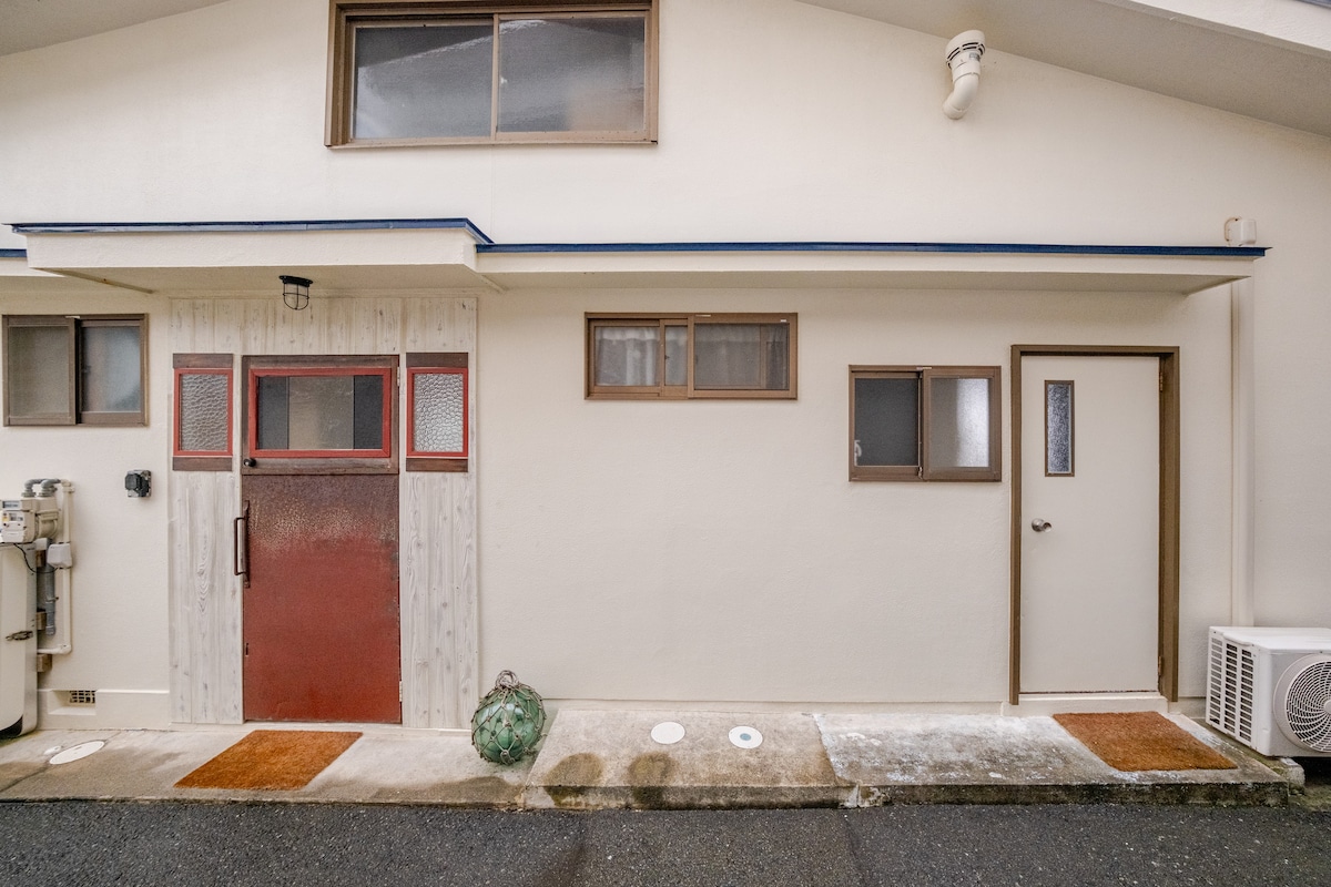 The exterior of the beach house displays a clean and modern aesthetic, featuring a combination of large windows and a distinctive red door. A smooth pathway leads to the entrance. Outdoor spaces include a small area with stepping stones and greenery.