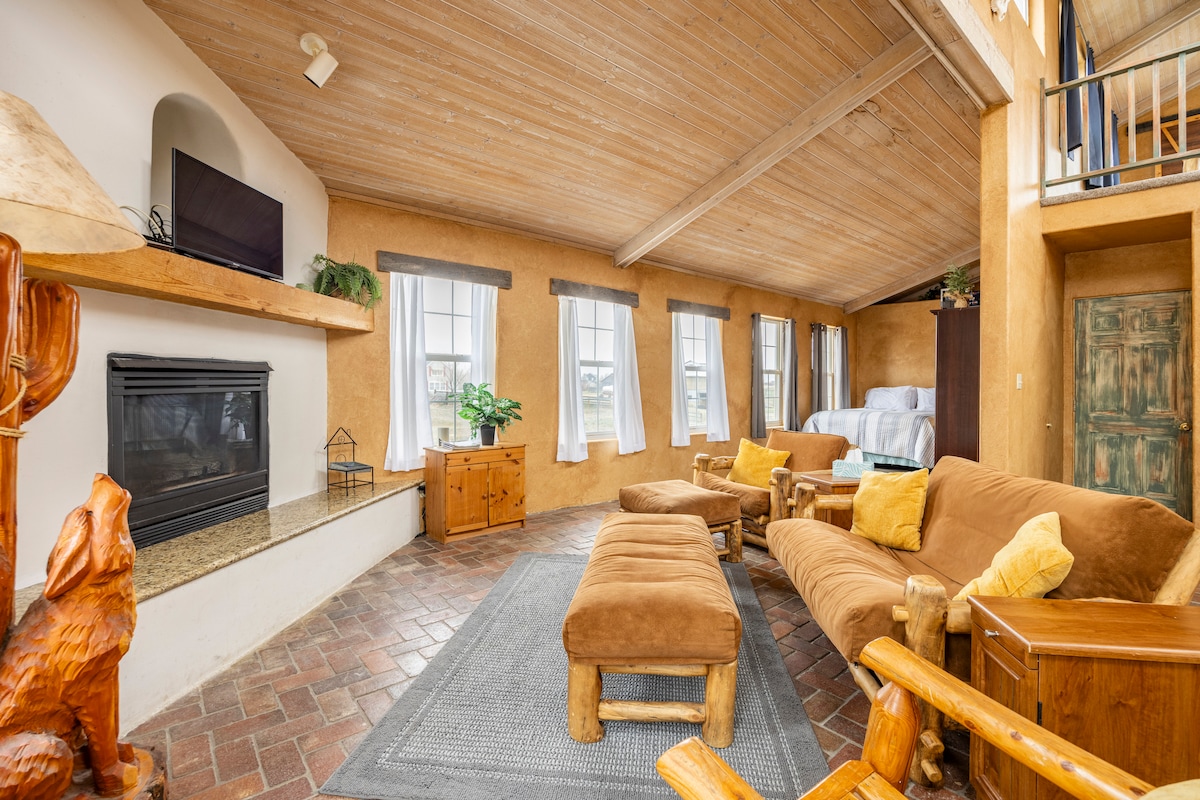 A spacious living area is characterized by warm-toned walls and a wood ceiling, featuring large windows that invite natural light. A cozy seating arrangement includes a sofa and upholstered chairs, while a fireplace offers a central focal point. Decorative wooden elements enhance the rustic charm.