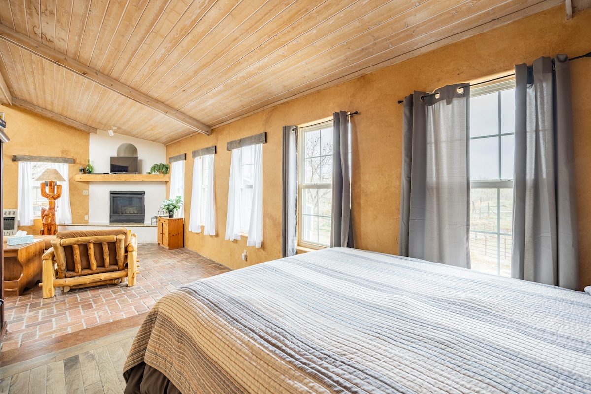 A spacious bedroom is featured with soft earth-toned walls and wooden ceiling beams. Natural light filters through multiple windows, which are dressed in simple curtains. A cozy seating area with a wooden sofa is visible alongside a brick fireplace, creating a relaxed environment.