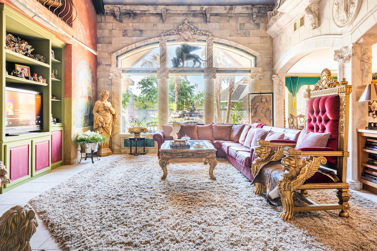 A spacious living area is characterized by ornate decor and plush seating. A large rug covers the floor, and natural light streams through expansive windows, revealing views of a garden. Decorative sculptures and detailed wall art add to the unique ambiance of the space.