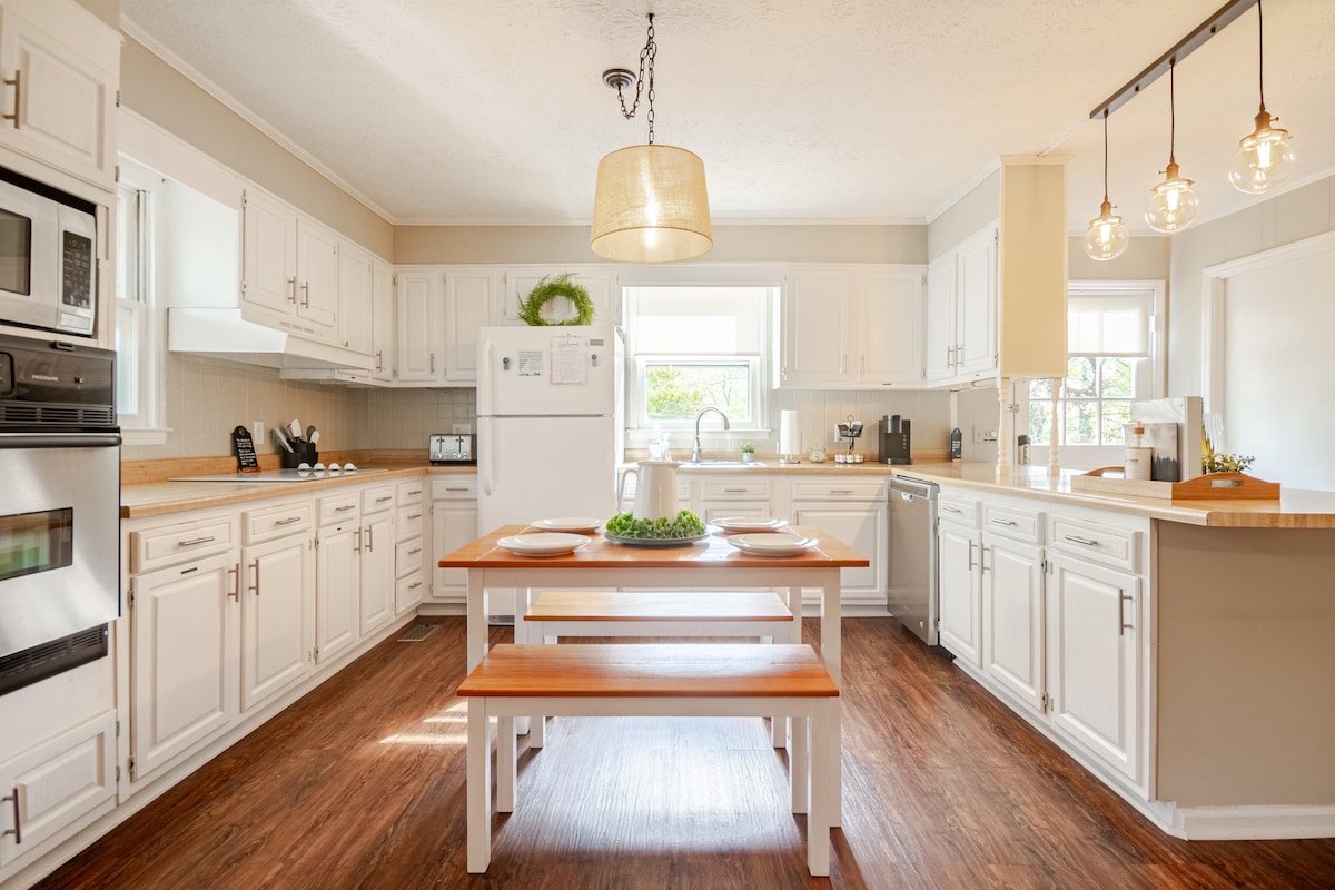 A well-equipped kitchen features white cabinetry, a spacious wooden table with benches, and modern appliances. Natural light floods the area through windows, highlighting the clean, organized layout. Pendant lights hang above the kitchen island, providing a warm ambiance for cooking and dining.