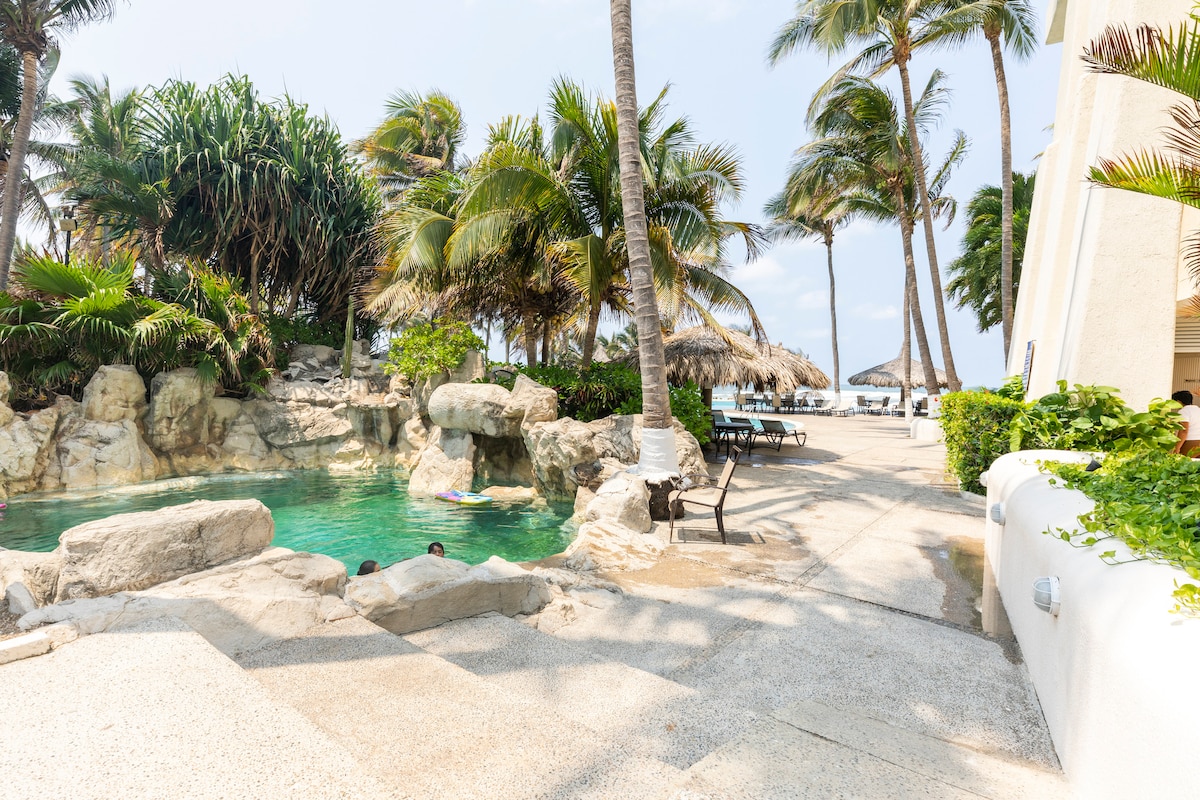 A natural stone swimming pool is framed by tropical palm trees, with a sunbathing area featuring lounge chairs. The path is lined with tropical greenery, leading towards the beach area beyond. Soft sunlight filters through the leaves, creating dappled light on the ground.